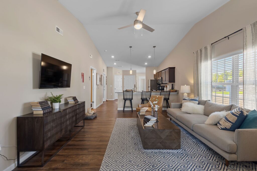 Modern open-concept living room and kitchen with neutral decor, a wall-mounted TV, a beige sofa, wood flooring, and large windows letting in natural light.