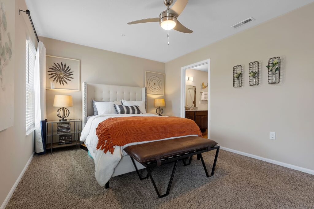 A neatly made bed with an orange throw blanket sits in a carpeted bedroom, featuring a ceiling fan, lamps, wall art, and an upholstered bench at the foot of the bed.