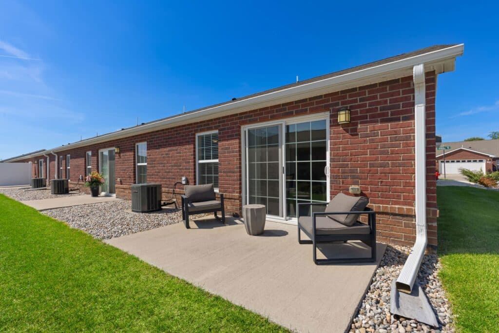 A brick single-story house with sliding glass doors opens onto a small concrete patio with outdoor chairs and potted plants, bordered by grass and decorative rocks.