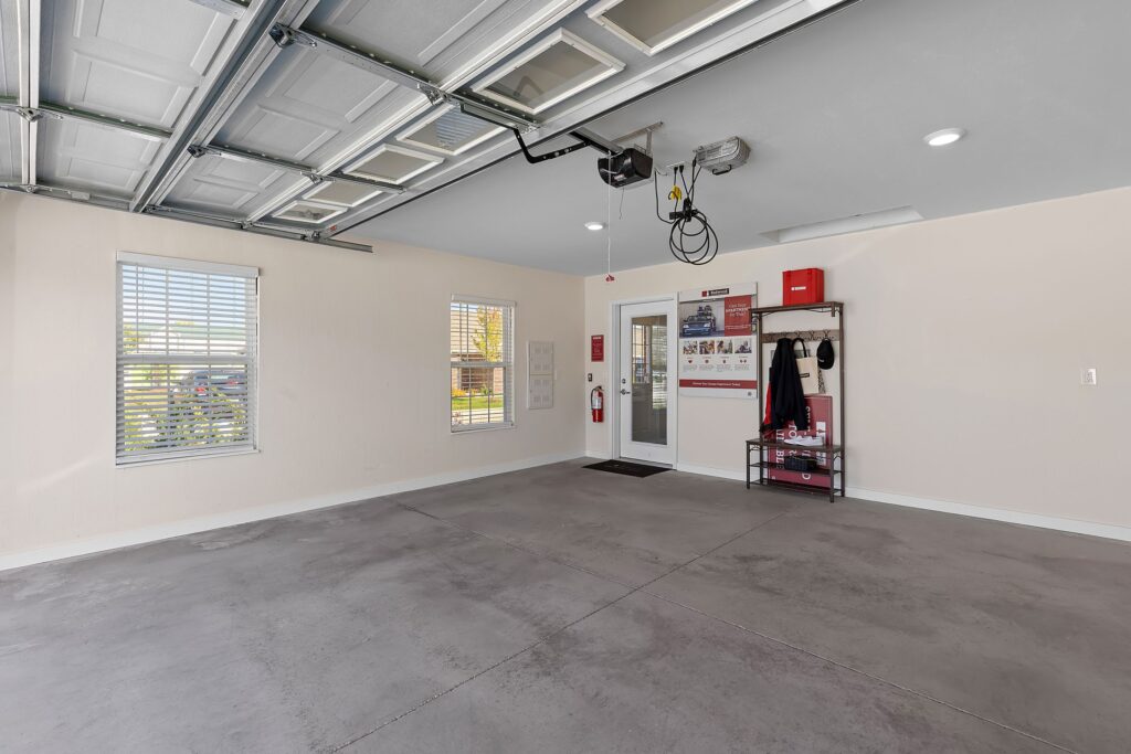 Spacious, clean garage with open door, gray concrete floor, beige walls, two windows, and a shelving unit near the entrance door.