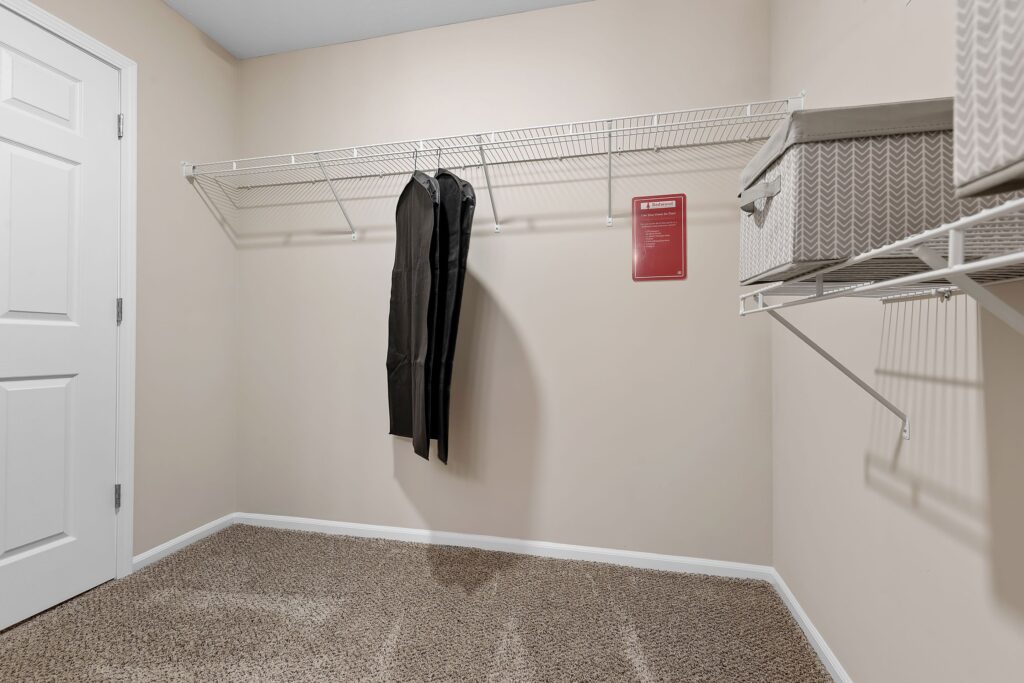 Empty walk-in closet with beige walls, carpeted floor, wire shelving on two walls, two pairs of pants hanging, and a storage box on the shelf.