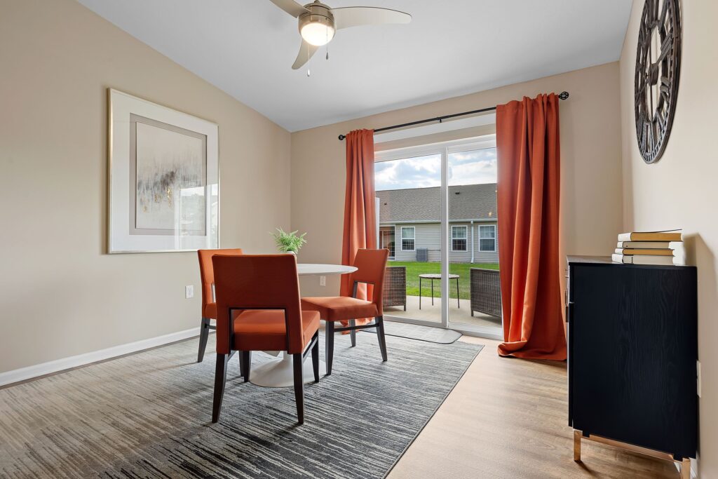 A small dining area with a round white table, two orange chairs, a black cabinet with books, orange curtains, and a large window overlooking a courtyard.