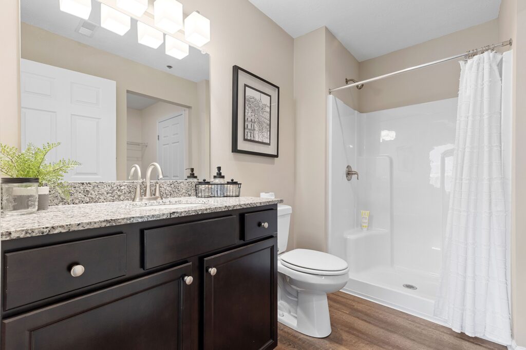 Modern bathroom with dark wood vanity, granite countertop, toilet, walk-in shower with white curtain, and a potted plant on the counter.