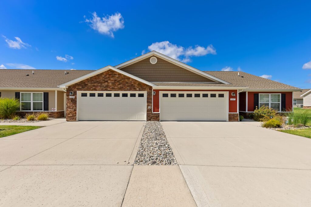 A duplex with two double garages, tan siding, brick accents, and a shared driveway divided by a strip of decorative rocks under a blue sky.