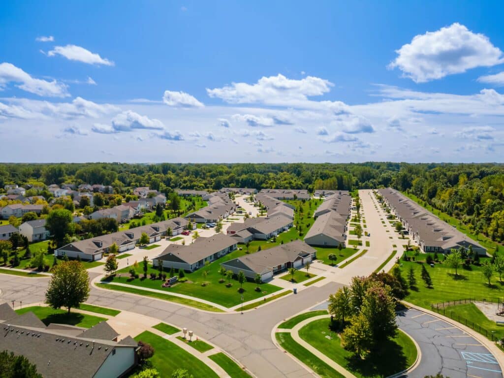 Aerial view of a suburban neighborhood with rows of single-family houses, green lawns, trees, and wide streets under a blue sky with scattered clouds.