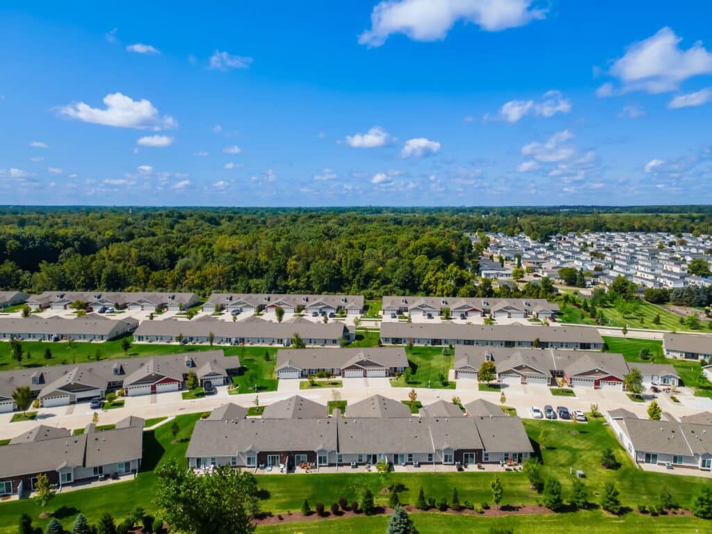 Aerial view of a suburban neighborhood with rows of single-story houses, green lawns, trees, and a forested area in the background under a partly cloudy sky.