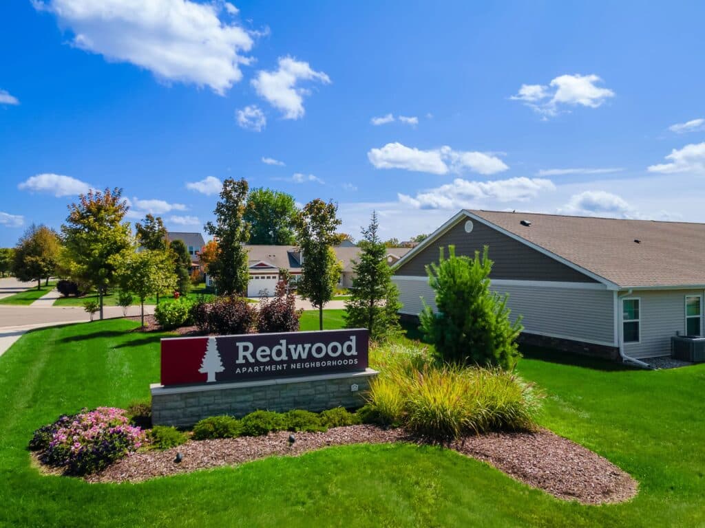 A landscaped entrance sign reading "Redwood Apartment Neighborhoods" in front of single-story homes on a sunny day.