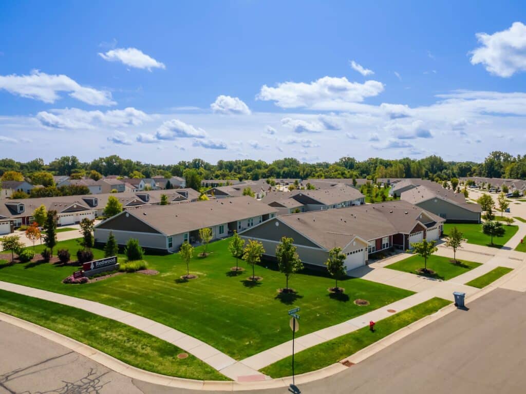 A suburban neighborhood with rows of single-story houses, well-maintained lawns, sidewalks, and trees under a blue sky with scattered clouds.