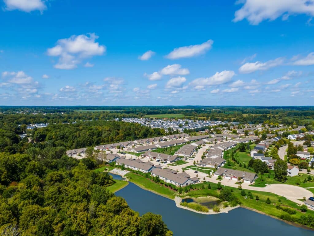 Aerial view of a suburban neighborhood with rows of houses, a small pond, trees, and open green fields under a blue sky with scattered clouds.