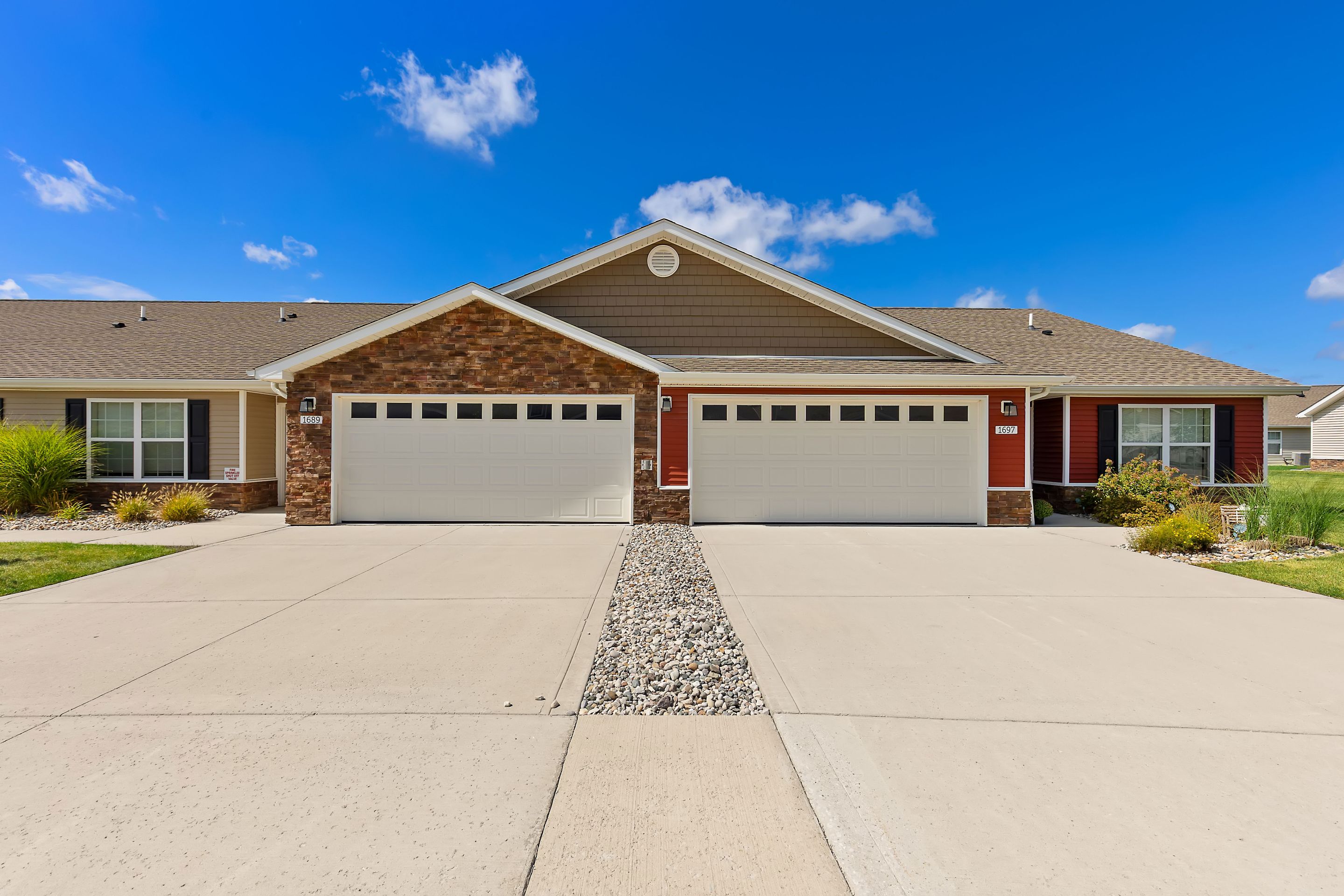 A duplex with two double garages, tan siding, brick accents, and a shared driveway divided by a strip of decorative rocks under a blue sky.