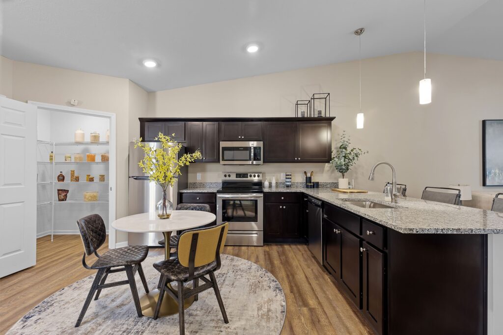 Modern kitchen with dark cabinets, stainless steel appliances, a round dining table with four chairs, granite countertops, and an open pantry in the background.