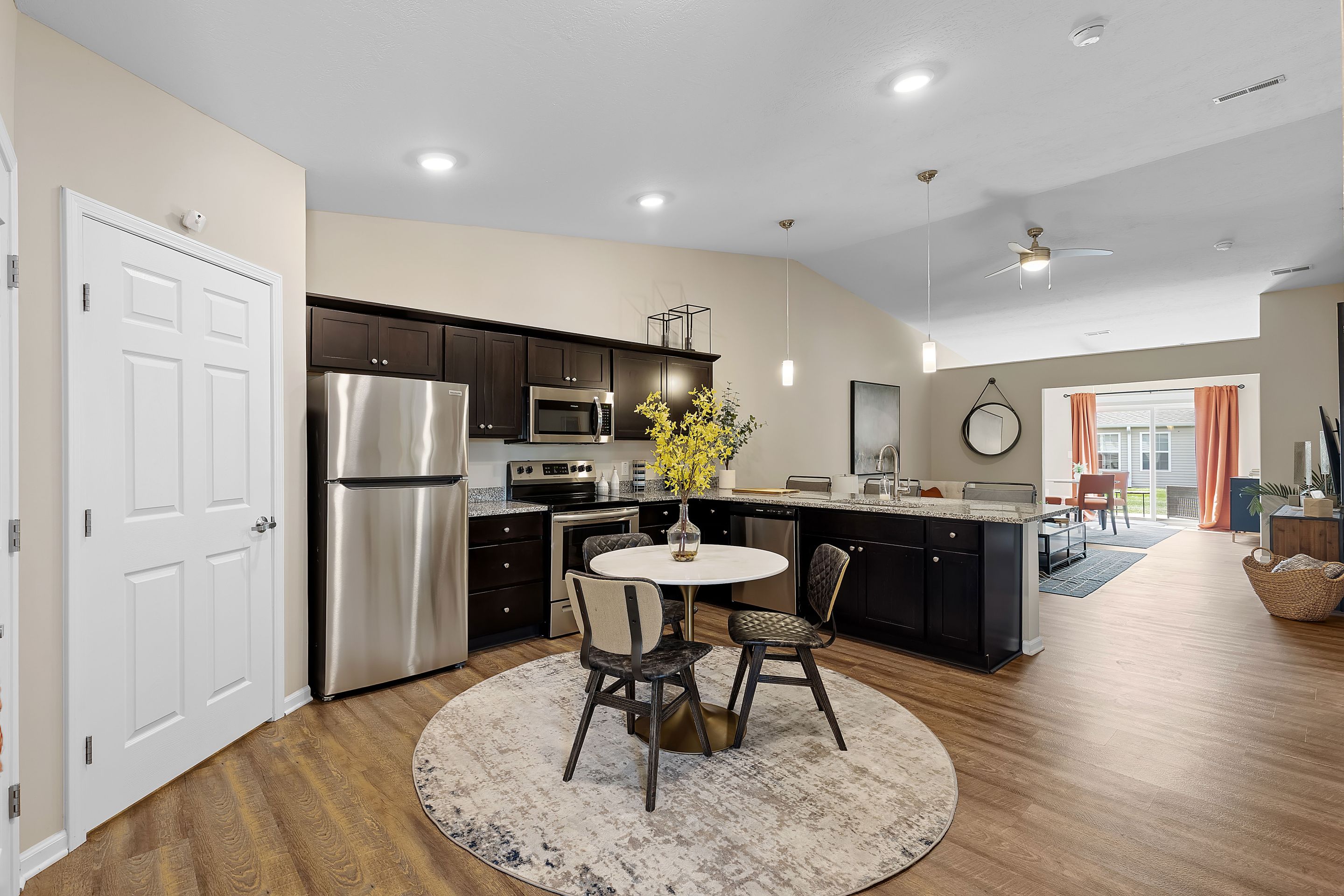 Modern open-concept kitchen and dining area with dark cabinets, stainless steel appliances, round table, and a living room visible in the background.