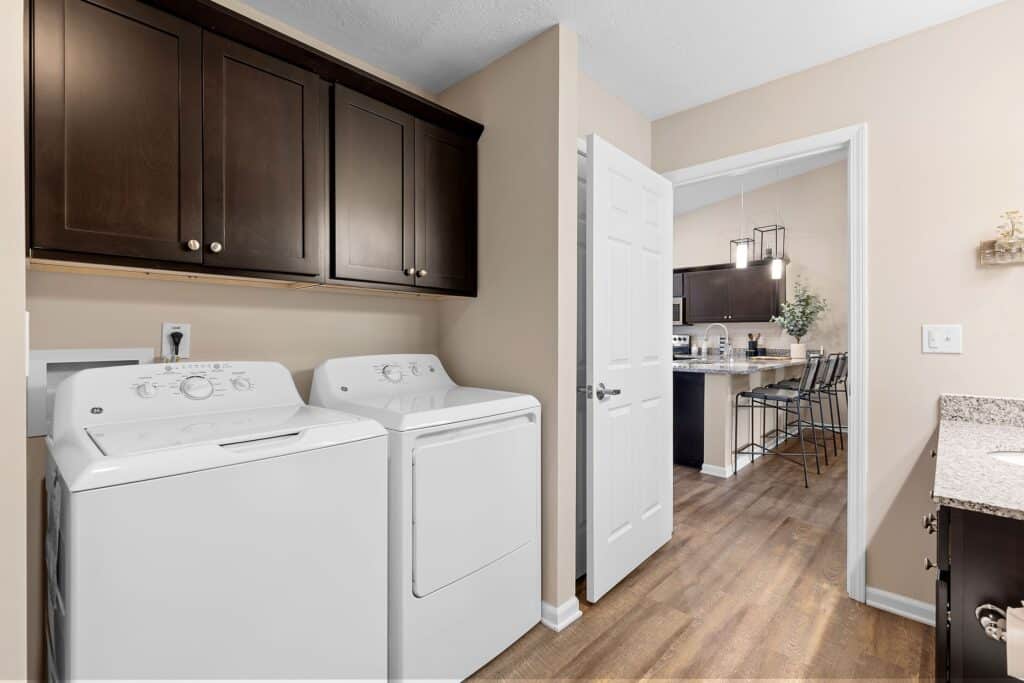 Laundry room with a white washing machine and dryer, dark wood cabinets above, and a view into a kitchen with bar stools and pendant lights.