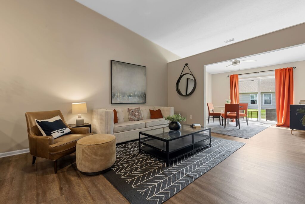 A modern living room with a beige sofa, patterned rug, black coffee table, and adjacent dining area with orange curtains and a window view.