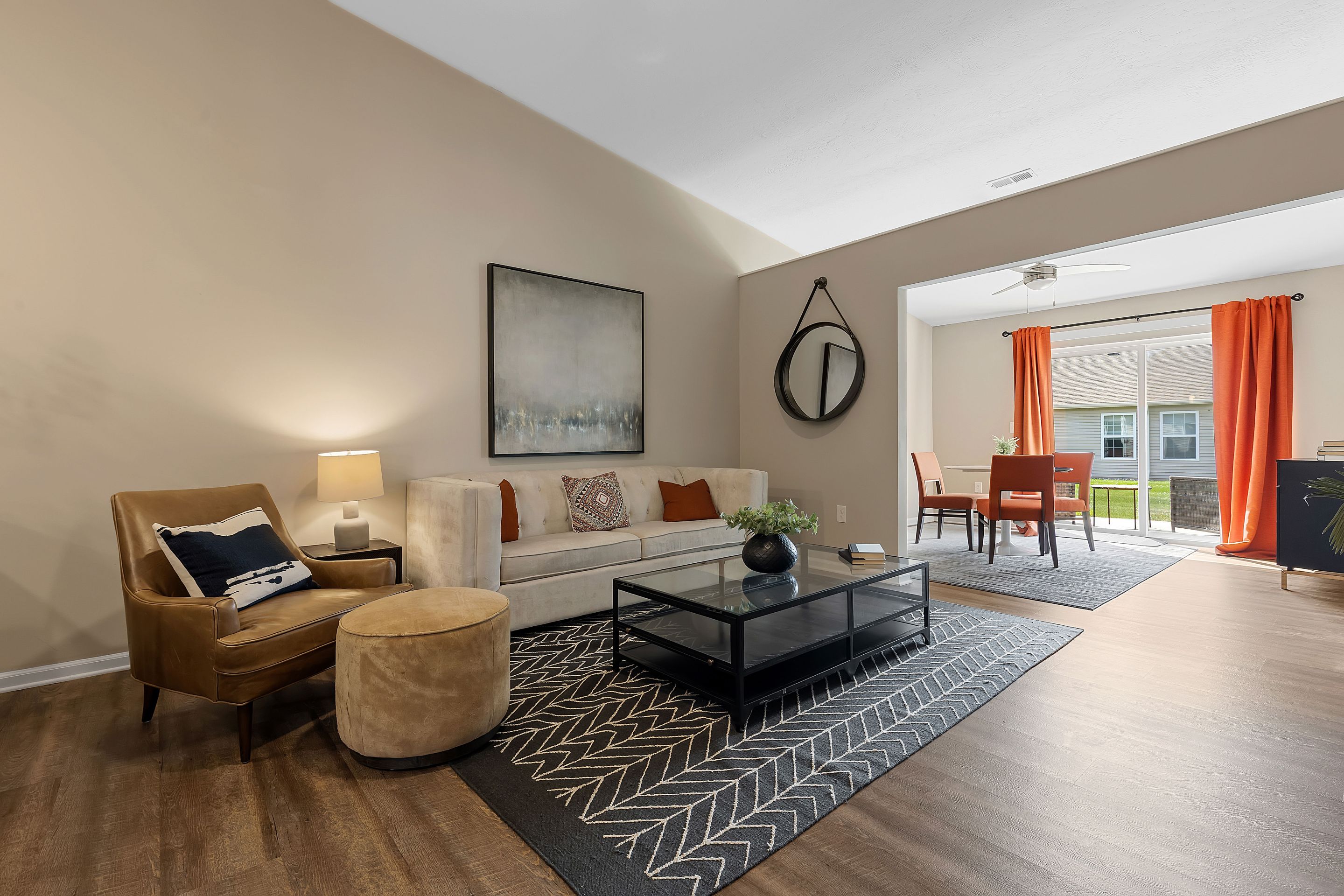 A modern living room with a beige sofa, patterned rug, black coffee table, and adjacent dining area with orange curtains and a window view.