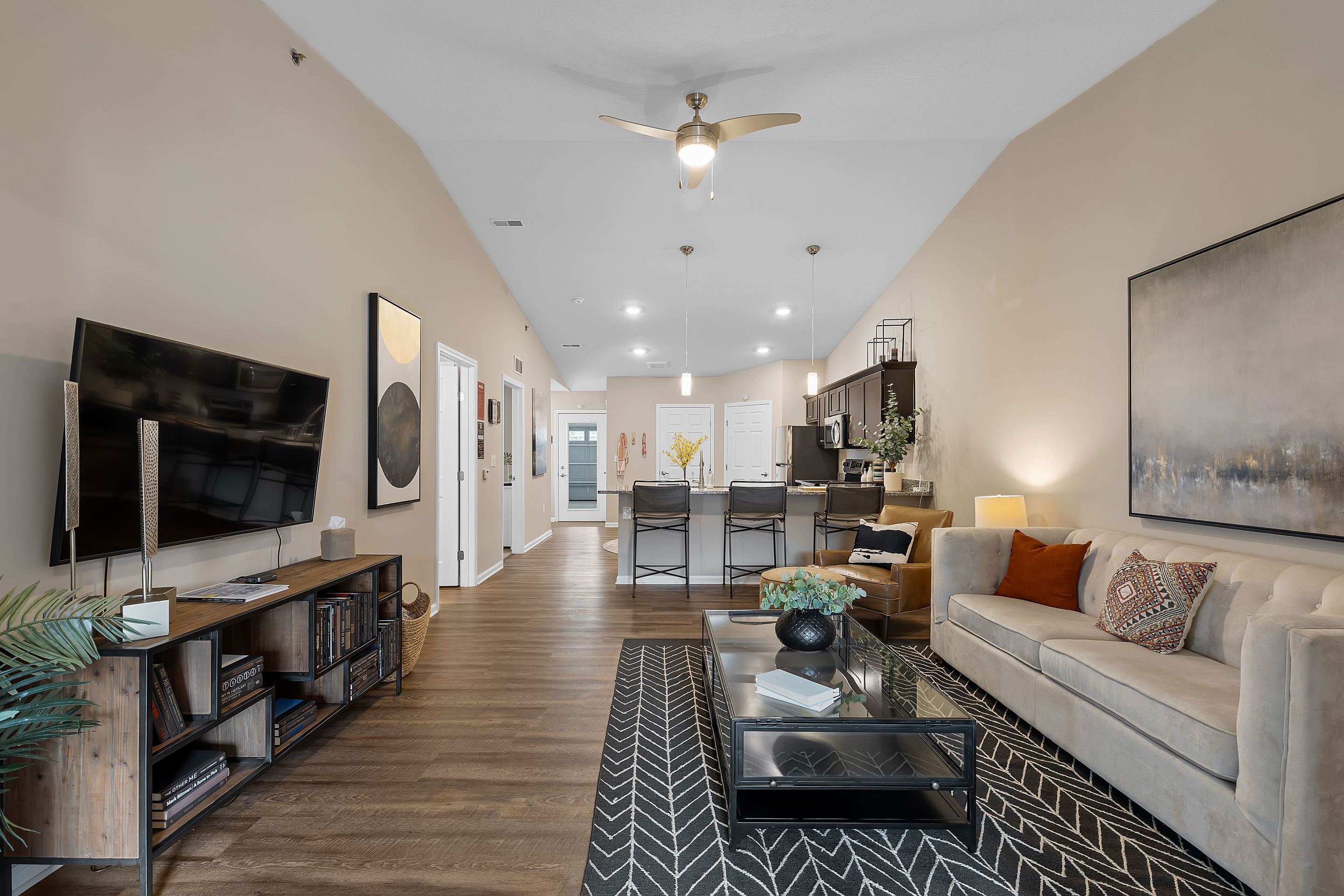 Modern open-plan living room and kitchen with beige walls, wood flooring, a beige sofa, glass coffee table, wall-mounted TV, and bar seating at the kitchen island.