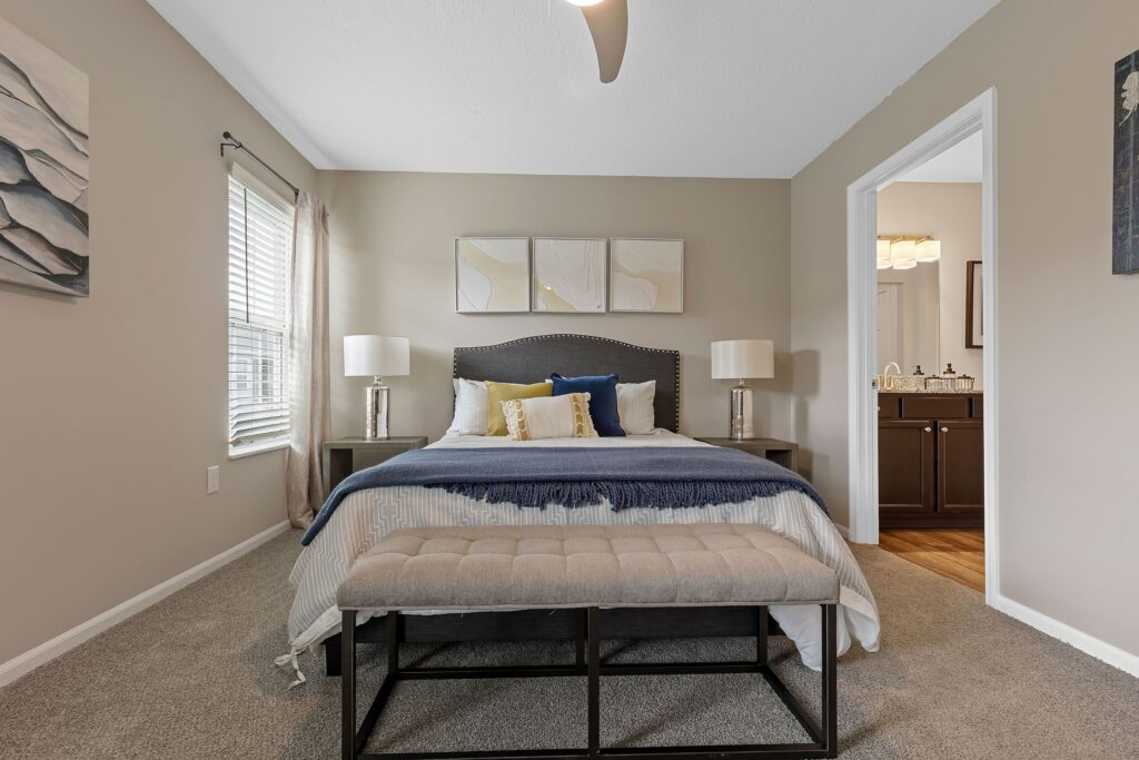 A neatly made bed with decorative pillows and a bench at the foot, two bedside tables with lamps, and a view into a bathroom in a neutral-toned bedroom.