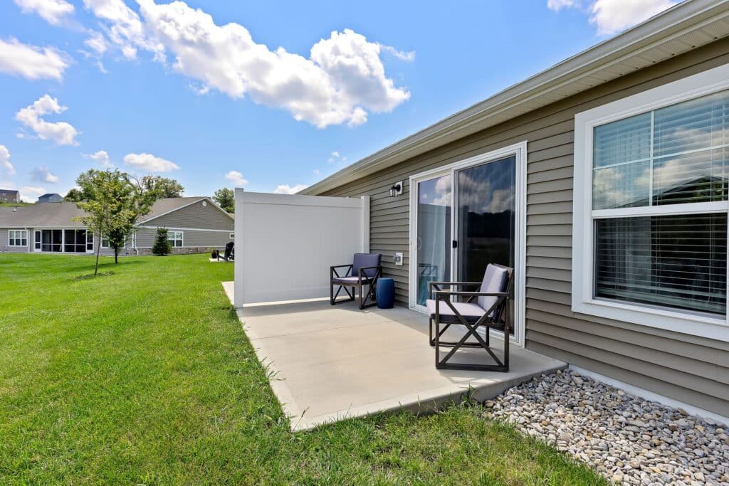 Concrete patio with two chairs, a small table, and privacy screen beside a beige house with sliding glass doors and large windows; grassy yard and neighboring homes visible.