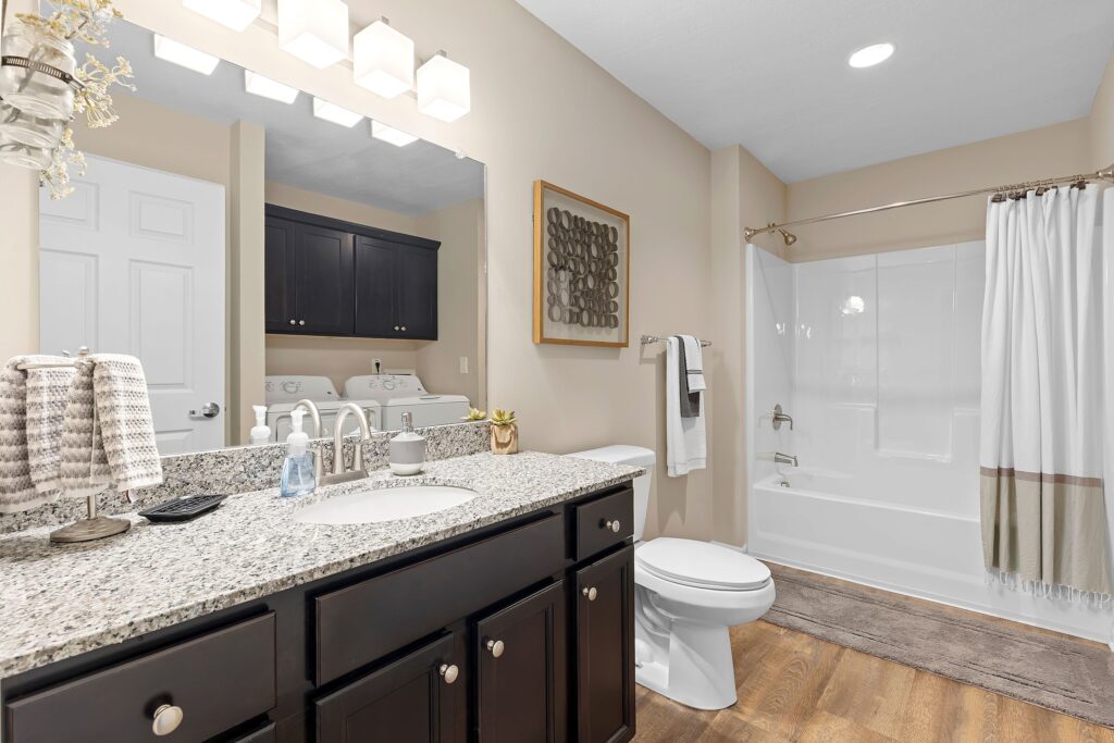 Modern bathroom with a granite countertop, double sinks, dark cabinets, a toilet, and a shower-tub combo with a white curtain; light wood floor and neutral walls.