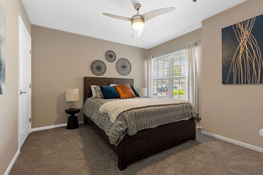 A modern bedroom features a dark wood bed with patterned bedding, a side table with a lamp, wall art above the bed, a ceiling fan, and a window with blinds and curtains.