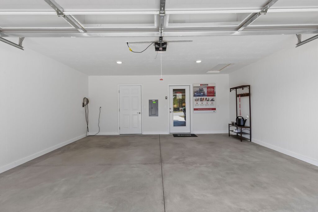Clean, empty two-car garage with white walls, concrete floor, a wall-mounted shelf, electrical panel, and a windowed door leading outside.