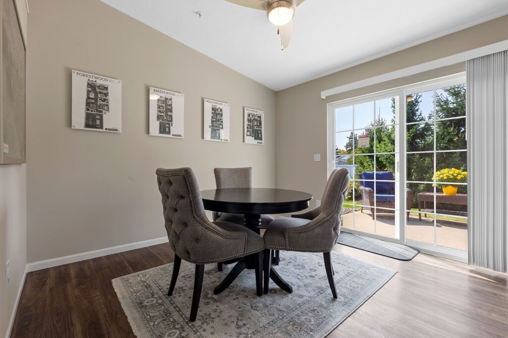 A dining area with a round black table, four gray upholstered chairs, wall art, wood flooring, and a sliding glass door opening to a patio with outdoor greenery.