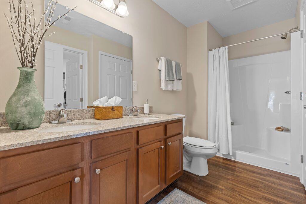 Modern bathroom with double-sink vanity, large mirror, wooden cabinets, toilet, shower with white curtain, and decorative vase on the countertop.