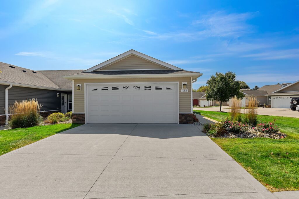 Single-story suburban house with a double garage, beige siding, white trim, and a concrete driveway, surrounded by green lawn and landscaping under a clear blue sky.