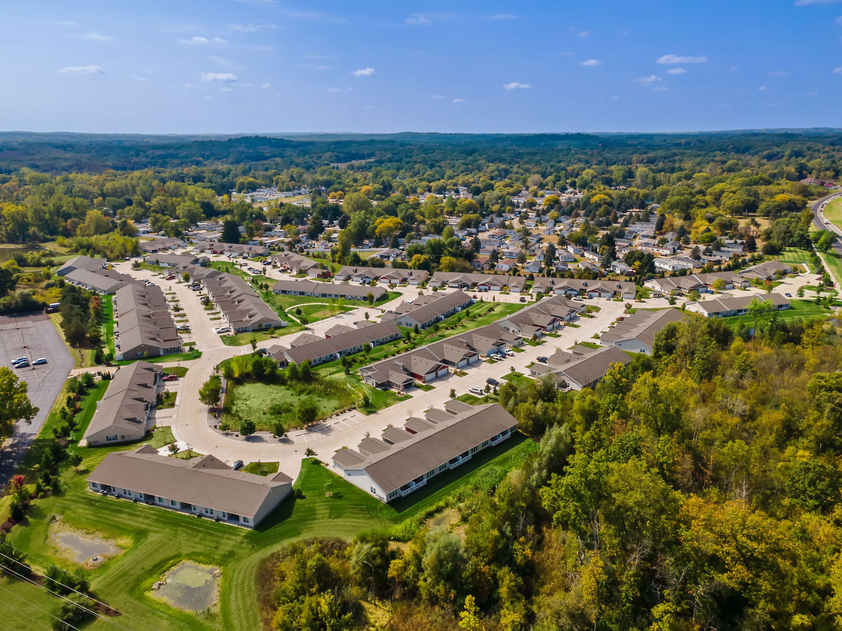Aerial view of a suburban neighborhood with rows of single-story houses, green lawns, trees, ponds, and a forested area in the background under a blue sky.