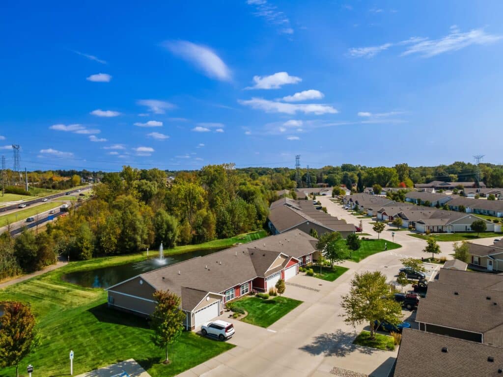 A suburban residential neighborhood with single-story houses, green lawns, trees, a small pond with a fountain, and a nearby highway under a blue sky.