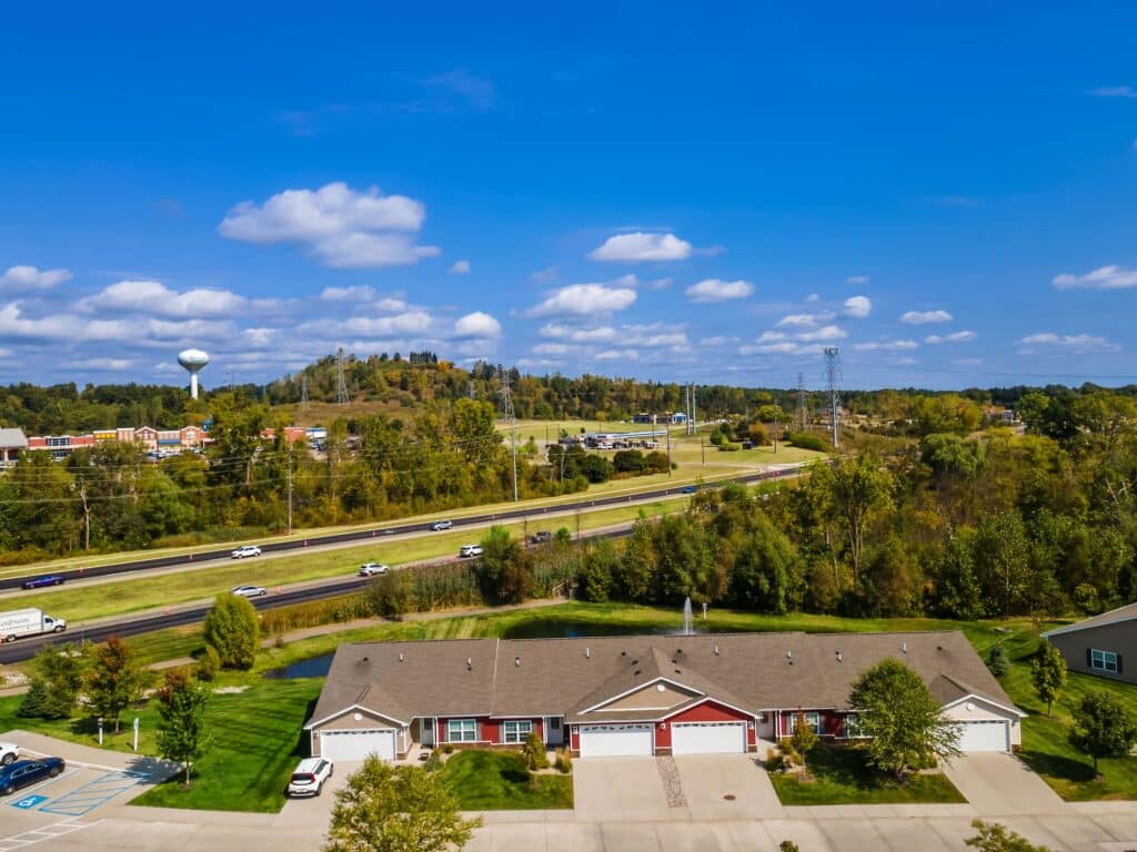 Aerial view of a suburban residential building with a green lawn, a parking lot, and a highway with cars passing by; trees and a water tower are visible in the background under a blue sky.
