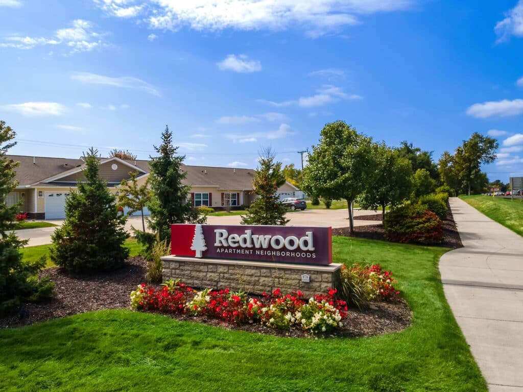Redwood Apartment Neighborhoods sign surrounded by flowers and shrubs, with residential buildings and a sidewalk in the background under a blue sky.