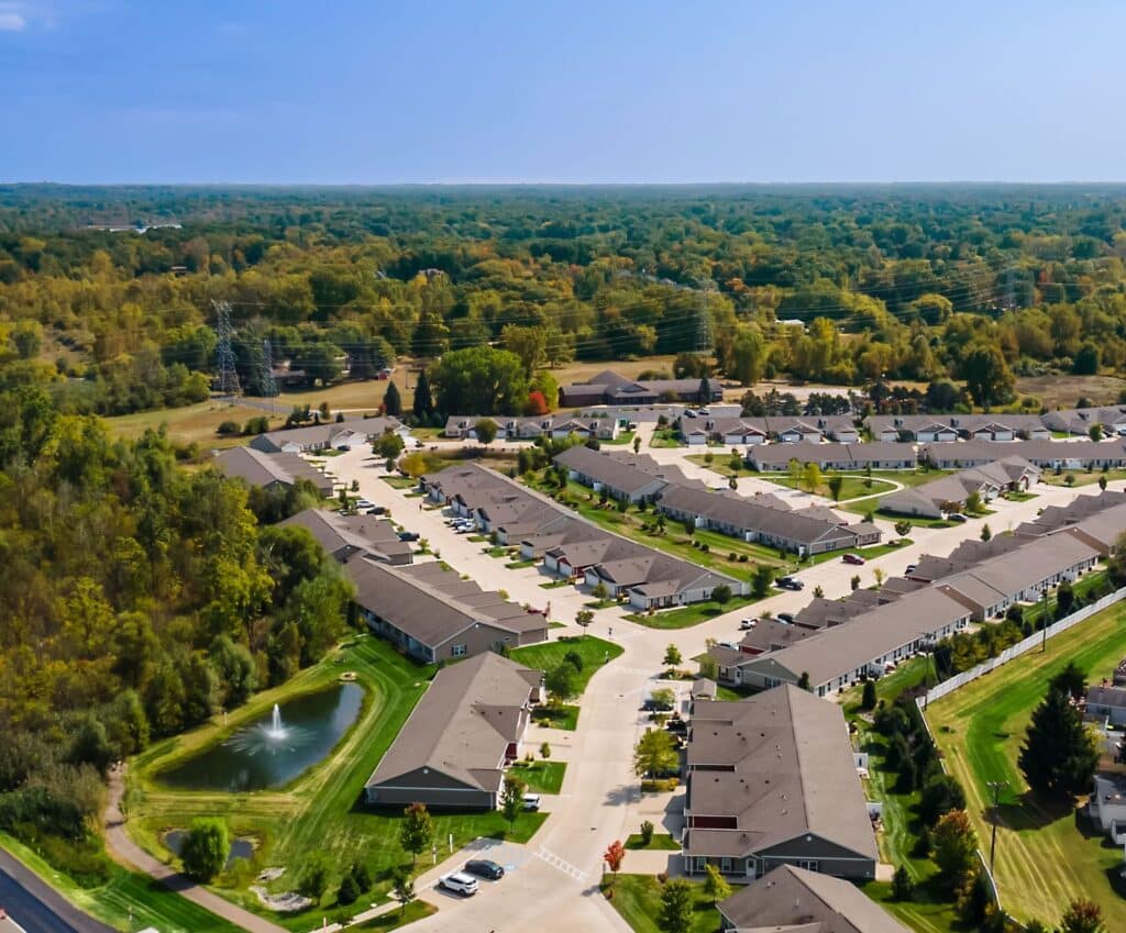 Aerial view of a suburban neighborhood with rows of houses, green lawns, a small pond with a fountain, and dense trees in the background.