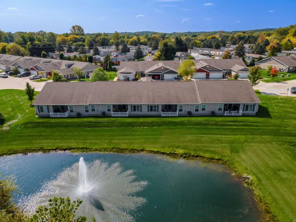 A row of single-story homes with porches faces a small pond with a fountain, surrounded by green grass and additional houses in the background.
