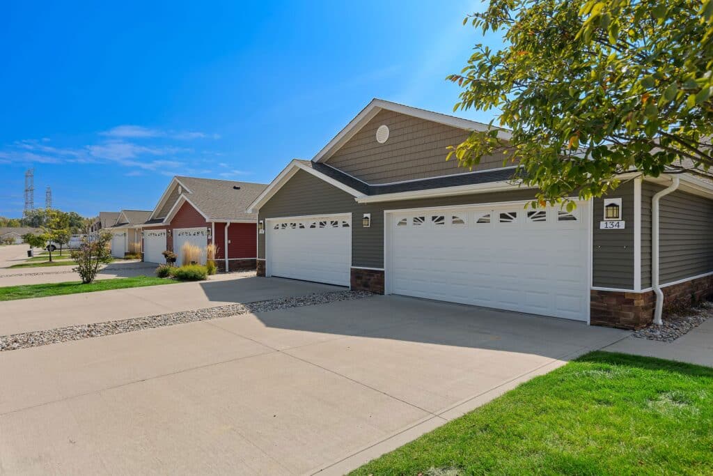 Row of single-story suburban houses with attached garages, concrete driveways, and manicured lawns on a clear, sunny day.