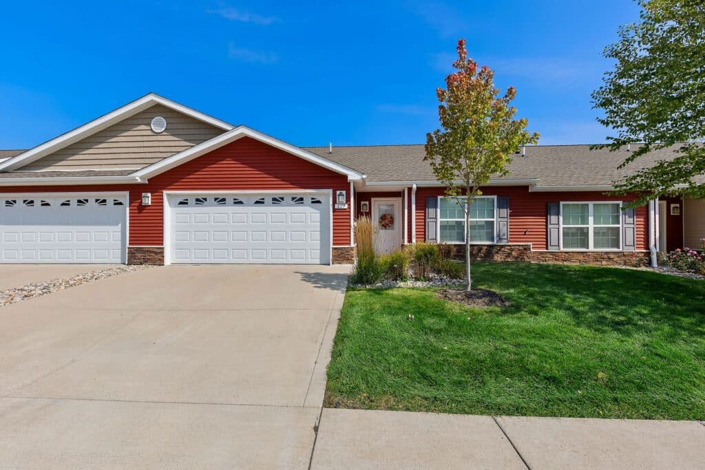 Single-story red duplex with white trim, two white garage doors, a small front yard with green grass, and a young tree by the front entrance.