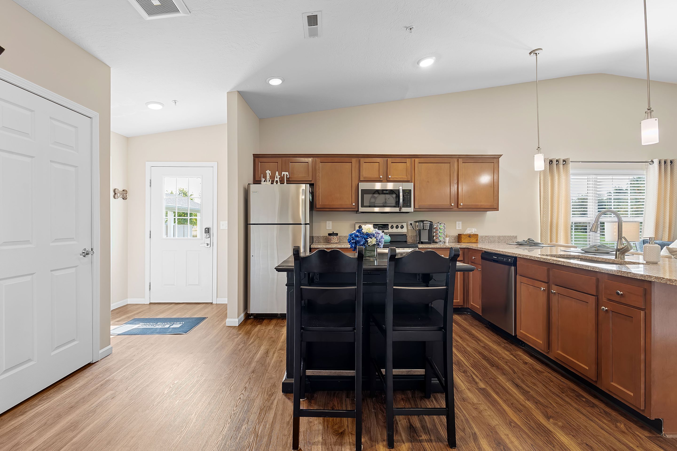 Modern kitchen with stainless steel appliances, wooden cabinets, a central island with two chairs, and hardwood flooring, featuring a window and door to the outside.