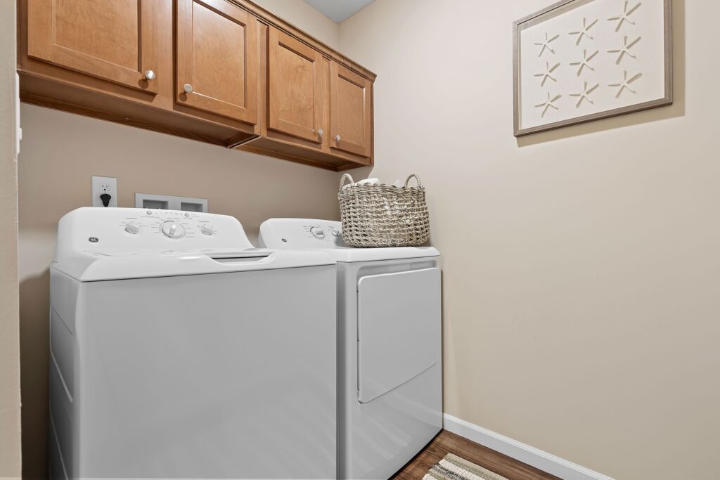 A laundry room with a white washing machine and dryer, wooden cabinets above, a woven basket on the dryer, and a framed artwork on the beige wall.