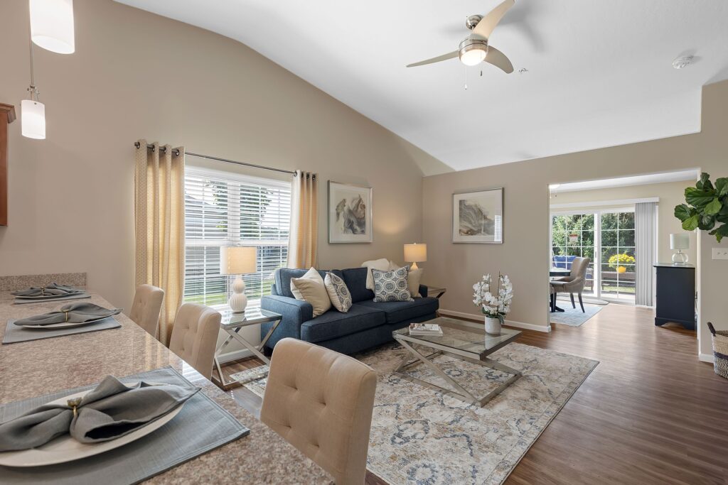 A modern living room with a blue sofa, coffee table, and patterned rug, adjacent to a dining counter and a sunlit room visible through double doors.