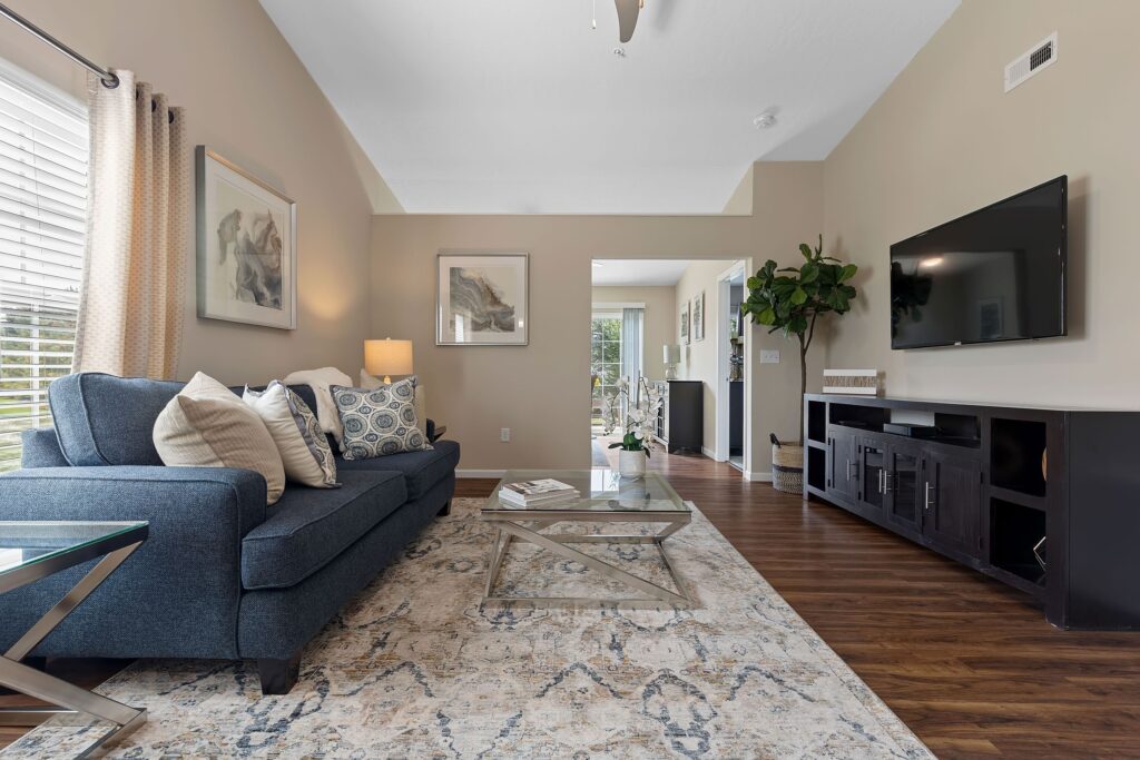 A modern living room with a blue sofa, patterned rug, glass coffee table, wall-mounted TV, and artwork on beige walls with wooden flooring.