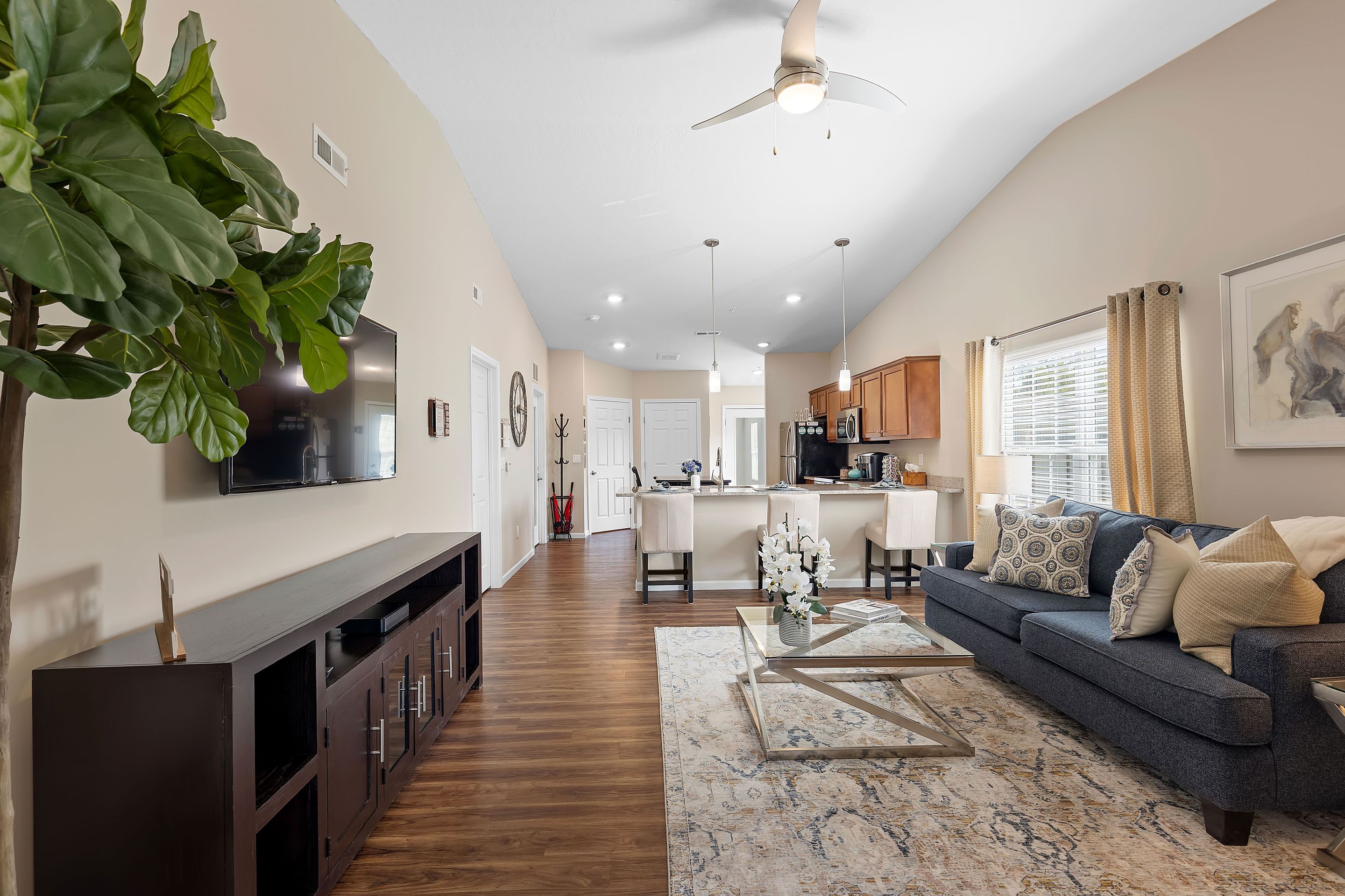 Modern living room with a blue sofa, patterned rug, wall-mounted TV, and open kitchen area with wooden cabinets and bar stools, lit by natural light from large windows.