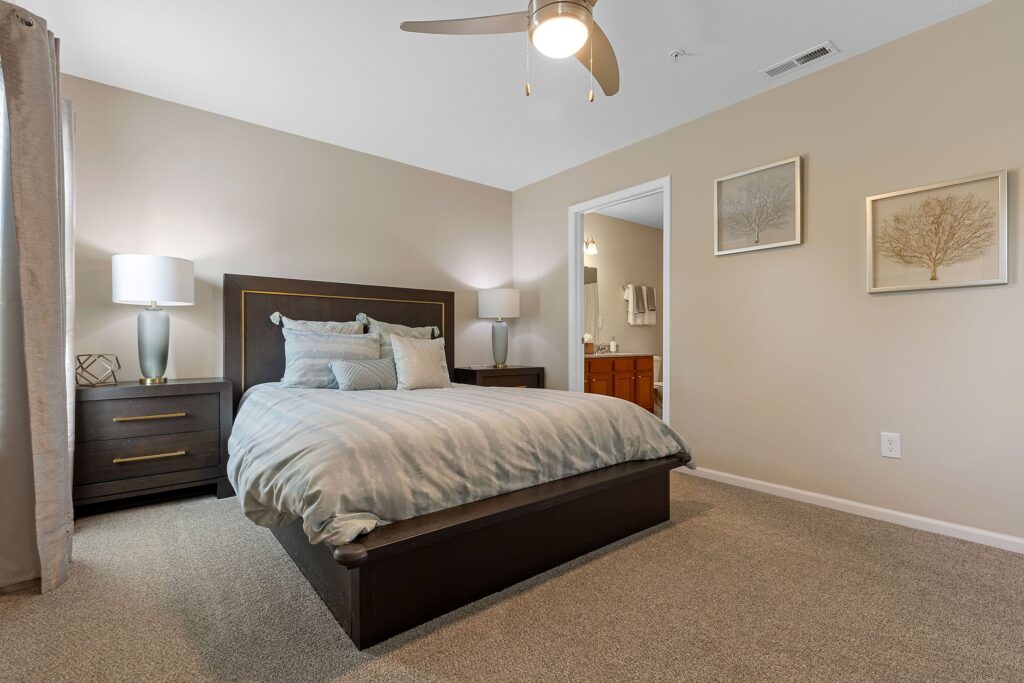 A neatly made bed with striped bedding in a beige bedroom, two nightstands with lamps, and an open door leading to a bathroom.