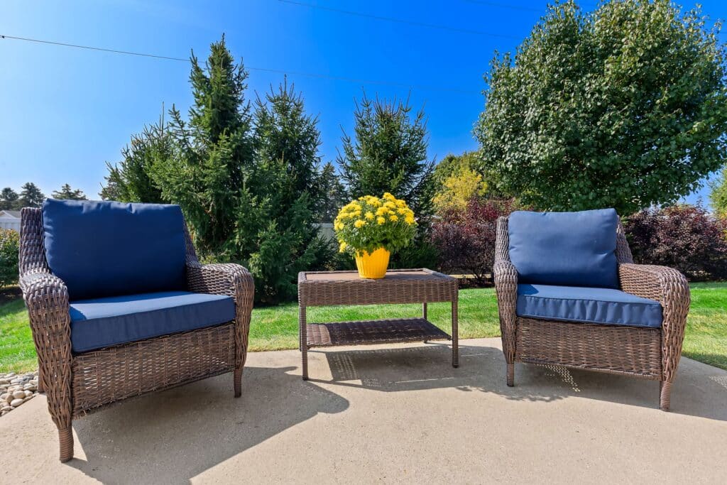 Two wicker patio chairs with blue cushions and a matching table with a yellow potted plant are set on a concrete patio in a sunny, landscaped backyard.