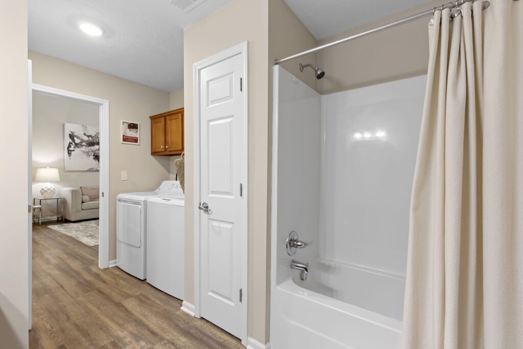 A bathroom with a white bathtub and shower curtain is next to a laundry area with a washer and dryer. The flooring is wood, and there is a door leading to another room.