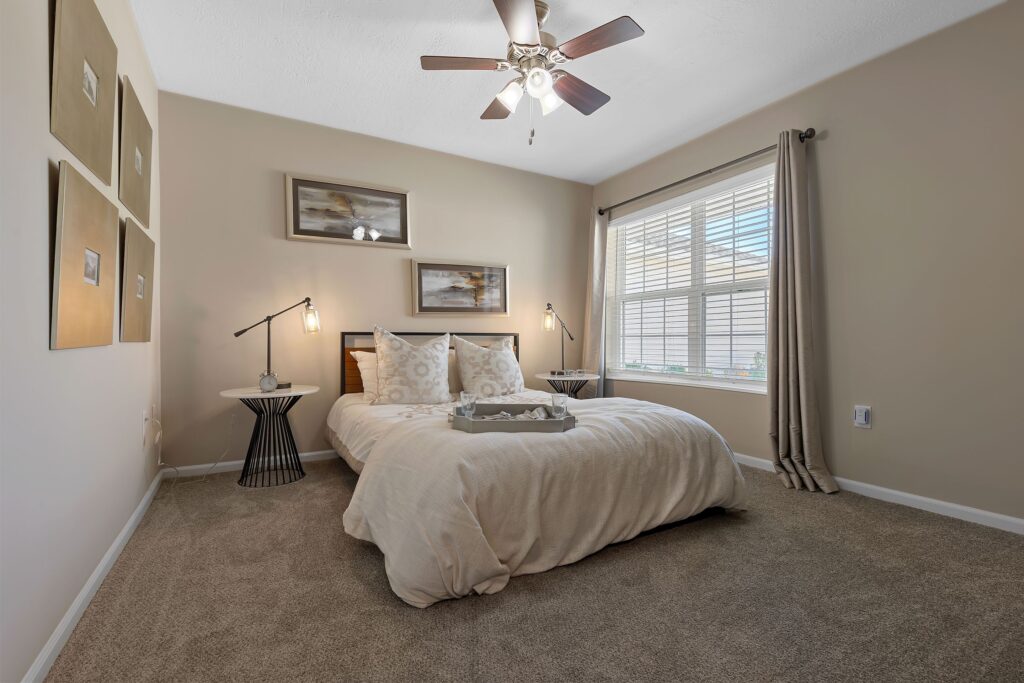 A modern bedroom with a neatly made bed, two side tables with lamps, wall art above the headboard, and a large window with beige curtains letting in natural light.