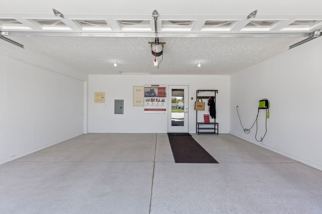 Open, clean two-car garage with white walls, a door and window at the back, wall-mounted electrical panels, coat rack, bench, and a charging device on the right wall.