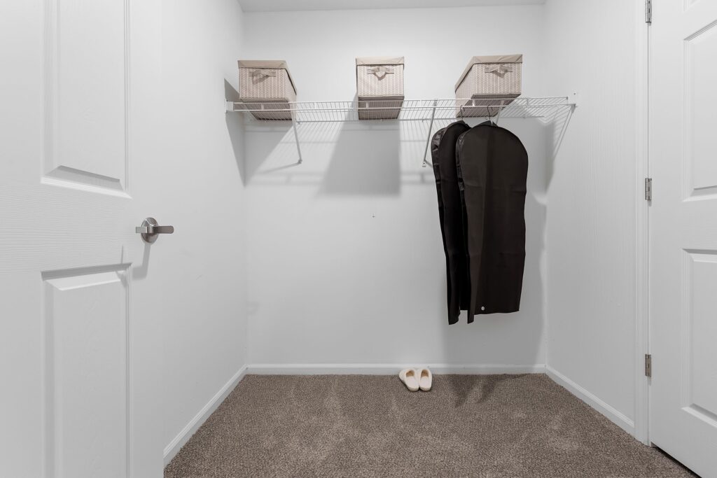 A clean, empty walk-in closet with beige carpet, three storage bins on a wire shelf, two garment bags hanging, and a pair of white slippers on the floor.
