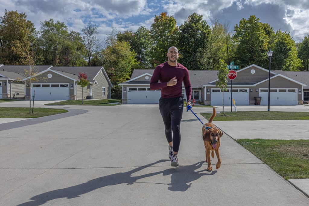A man jogging on a suburban street with a brown dog on a leash, with houses and trees in the background under a partly cloudy sky.