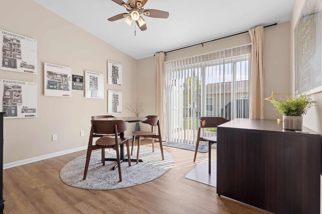 A small dining area with a round table and four chairs sits on a rug near sliding glass doors with vertical blinds, allowing natural light into the room.