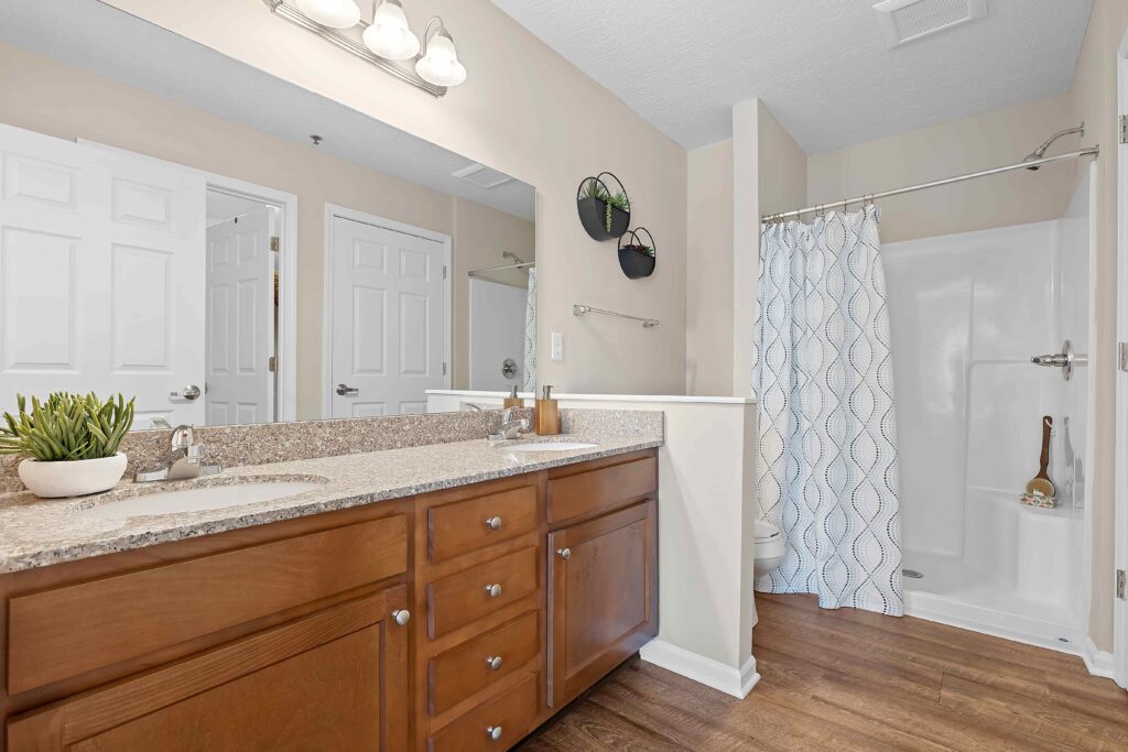 Bathroom with wooden vanity, granite countertop, potted plant, wall mirror, shower with white curtain, and beige walls.